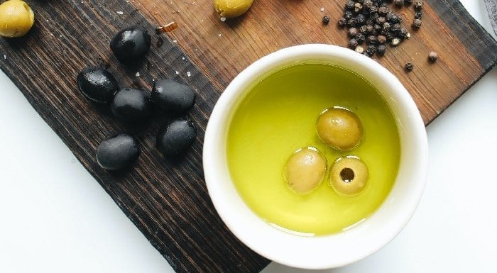 Bowl of olive oil with green olives, next to black olives and peppercorns