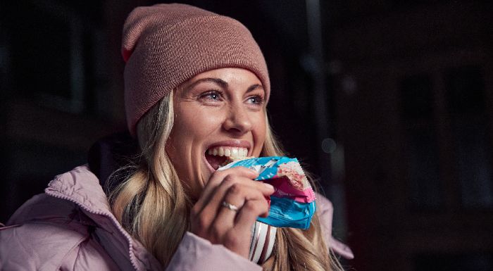 Woman in a beanie and puffer jacket smiling as she eats a snack bar