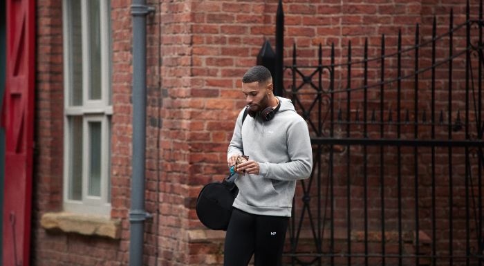 Man in a grey hoodie with a gym bag, checking his watch outside a brick building
