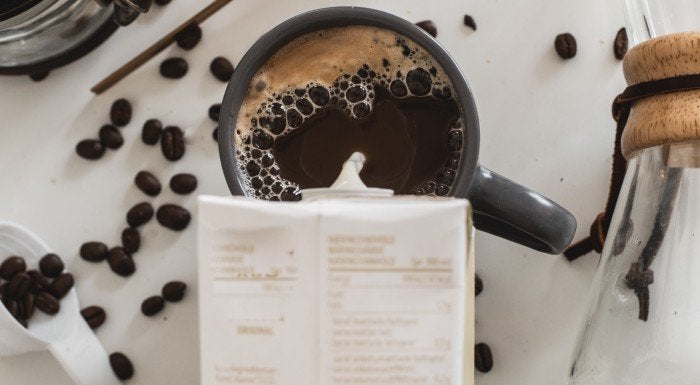 Carton of oat milk being poured into a cup of coffee, surrounded by coffee beans