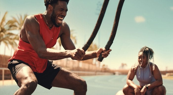 Man training with battle ropes outdoors while a woman watches
